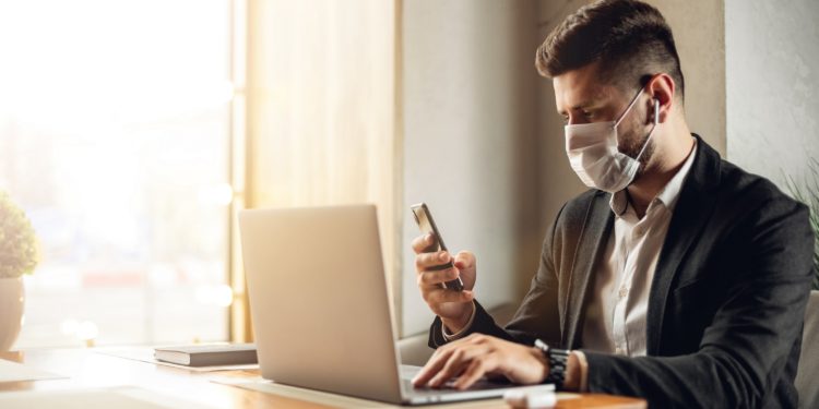 Mann mit Mundschutz sitzt vor einem Laptop und hält sein Smartphone in der rechten Hand