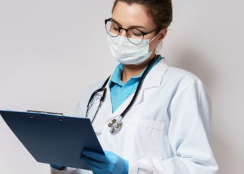 Ärztin mit Brille, weißem Kittel, Mundschutz, Handschuhen und Stethoskop notiert etwas
