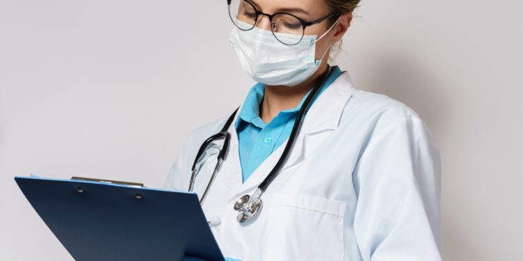 Ärztin mit Brille, weißem Kittel, Mundschutz, Handschuhen und Stethoskop notiert etwas