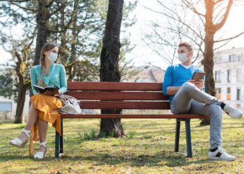 Mann und Frau mit Mund-Nasen-Schutz sitzen mit Abstand voneinander auf einer Parkbank