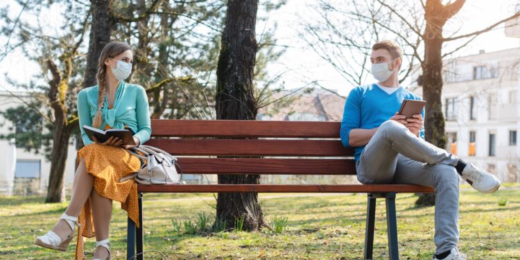 Mann und Frau mit Mund-Nasen-Schutz sitzen mit Abstand voneinander auf einer Parkbank