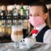 Eine weibliche Bedienung mit Mund-Nasen-Schutz serviert Kaffee