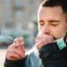Mann mit Zigarette und umgehängten Mund-Nasen-Schutz hustet in seine Hand