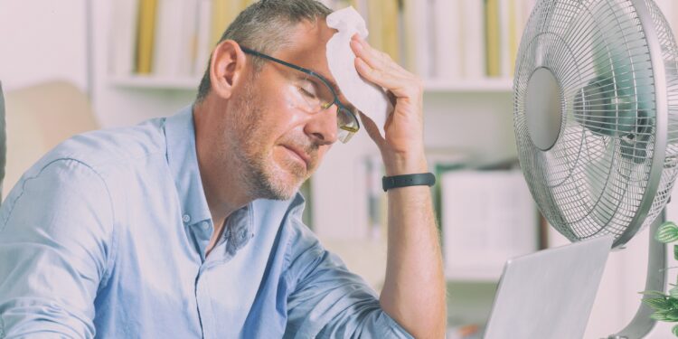 Mann im Homeoffice leidet unter Hitze und versucht, sich durch einen Ventilator abzukühlen