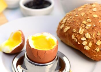 Ein geöffnetes Frühstücksei im Becher neben einem Brötchen und einer Tasse Kaffee