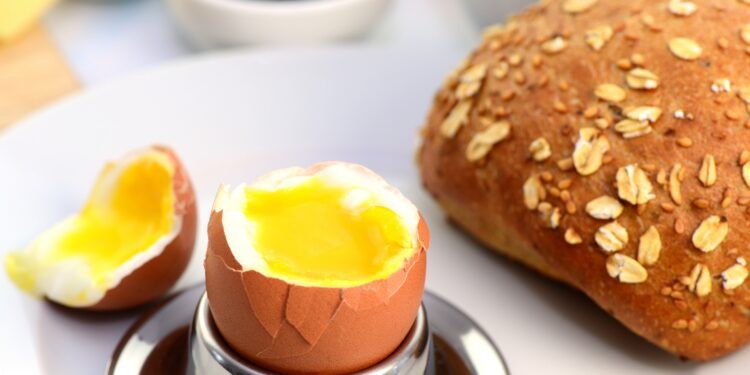 Ein geöffnetes Frühstücksei im Becher neben einem Brötchen und einer Tasse Kaffee