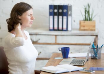 Junge Frau am Büroarbeitsplatz fasst sich an den schmerzenden Nacken