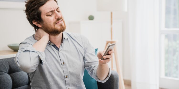 Mann mit schmerzverzerrtem Gesicht fasst sich an den Nacken und blickt auf sein Smartphone