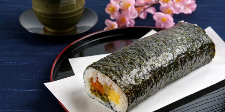 Eine Sushi-Rolle auf einem Teller vor einer Tasse mit Tee und Blumen auf einem Tisch