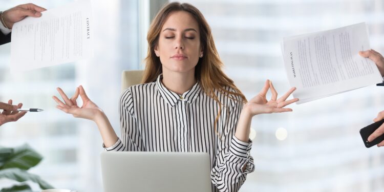 Junge Frau sitzt vor einem Laptop und macht eine Meditationsübung, während von allen Seiten Aufgaben an sie herangetragen werden