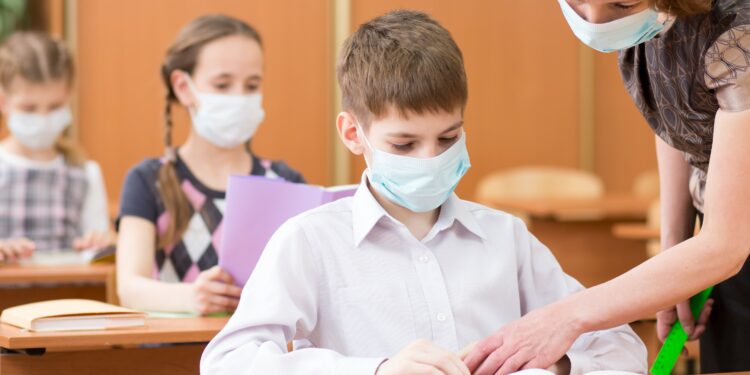 Schulkinder und Lehrerin mit Mund-Nasen-Bedeckungen im Klassenzimmer