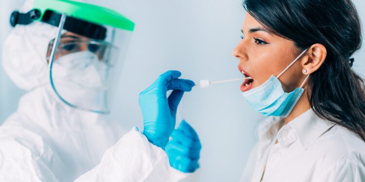 Medizinische Fachkraft in Schutzkleidung nimmt bei einer jungen Frau einen Rachenabstrich für einen Corona-Test
