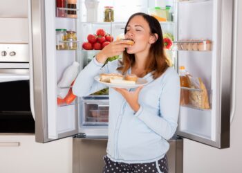 Frau mit Teller voller Kuchen in der Hand isst einen Donut vor dem geöffneten Kühlschrank