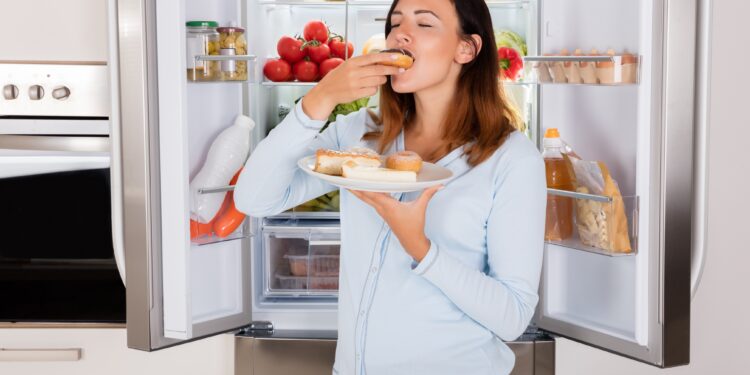Frau mit Teller voller Kuchen in der Hand isst einen Donut vor dem geöffneten Kühlschrank