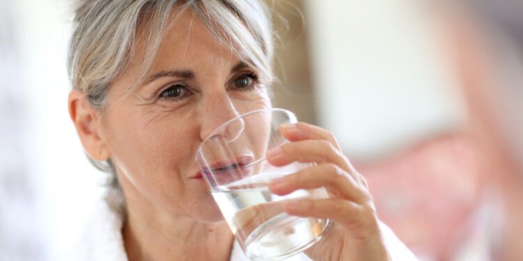 Eien Seniorin im Morgenmantel trinkt ein Glas Wasser