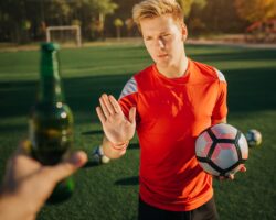 Ein Fußballer mit eine Fußball in der Hand weist eine angebotene Flasche Bier zurück