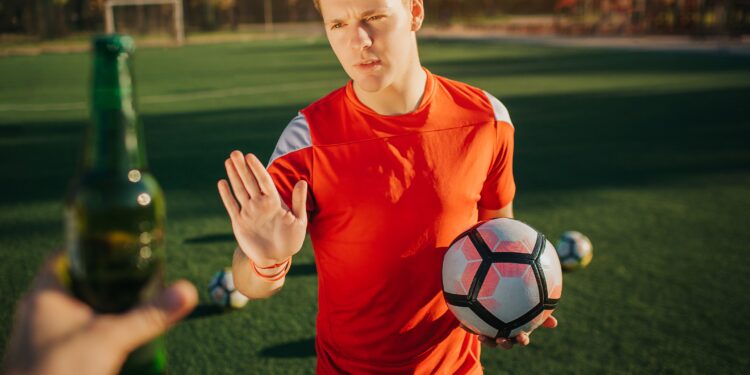 Ein Fußballer mit eine Fußball in der Hand weist eine angebotene Flasche Bier zurück