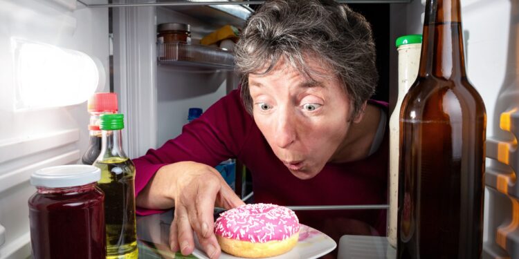 Eine Frau greift nach einem Donat im Kühlschrank.