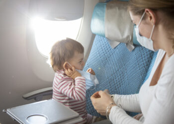 Frau mit Mund-Nasen-Schutz sitzt neben ihrem Kind im Flugzeug
