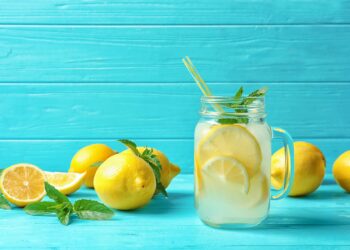 Ein Glas mit Zitronenwasser und einem Strohhalm und mehrere frische Zitronen auf einem Holztisch