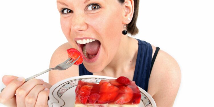 Frau mit weit geöffnetem Mund hält einen Teller mit einem Stück Erdbeerkuchen