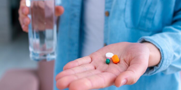 Mann hält in der linken Handfläche drei Tabletten, in der rechten Hand ein Glas Wasser