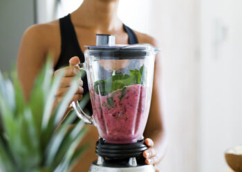 Junge Frau macht sich im Mixer einen Smoothie mit Spinat, Beeren, Bananen, Mandeln und Milch