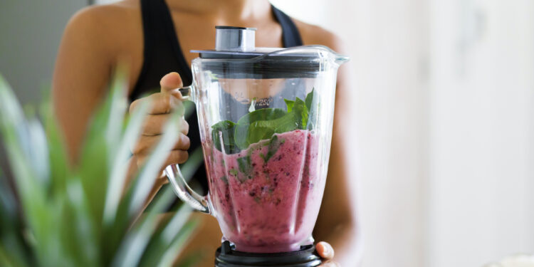 Junge Frau macht sich im Mixer einen Smoothie mit Spinat, Beeren, Bananen, Mandeln und Milch