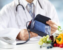 Obst und Gemüse und ein Maßband auf einem Tisch vor einem Arzt mit einem Blutdruckmessgerät