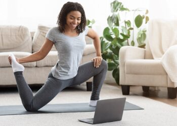 Frau trainiert im Wohnzimmer vor dem Laptop auf Yogamatte