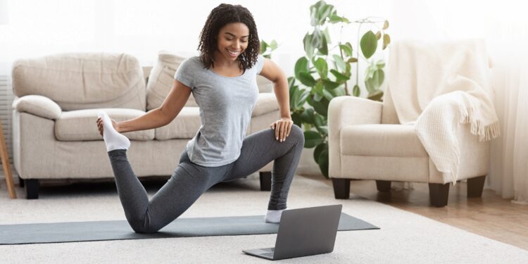 Frau trainiert im Wohnzimmer vor dem Laptop auf Yogamatte