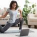 Frau trainiert im Wohnzimmer vor dem Laptop auf Yogamatte