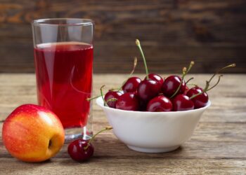 Ein Apfel, eine Schale mit Kirschen und ein Glas mit Saft stehen auf einer hölzernen Oberfläche.