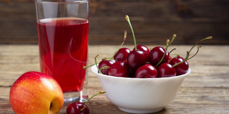 Ein Apfel, eine Schale mit Kirschen und ein Glas mit Saft stehen auf einer hölzernen Oberfläche.