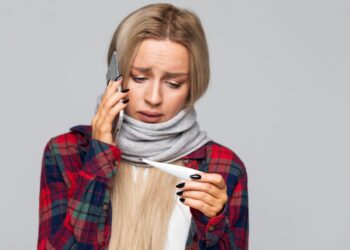 Junge Frau mit Erkältung blickt auf ein Fieberthermometer während sie mit einem Smartphone telefoniert