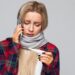 Junge Frau mit Erkältung blickt auf ein Fieberthermometer während sie mit einem Smartphone telefoniert