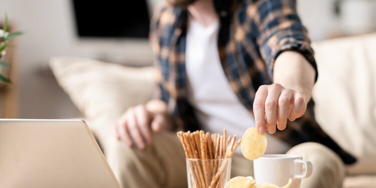 Mann sitzt am Laptop und greift zu Chips