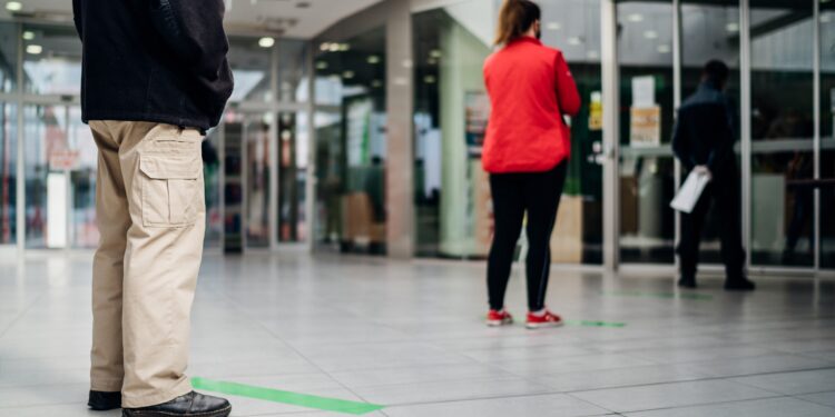 Mehrere Menschen stehen mit Abstand voneinander in einer Reihe vor einer Bank