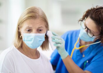 Ärztin mit Maske misst bei einer Frau kit Maske das Fieber im Ohr