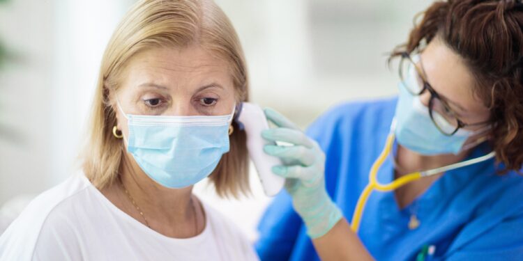 Ärztin mit Maske misst bei einer Frau kit Maske das Fieber im Ohr