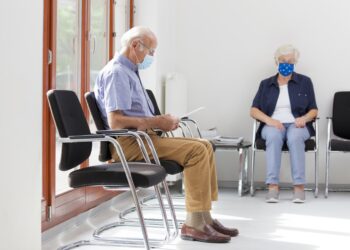 Eine Seniorin und ein Senior mit Mund-Nasen-Bedeckung sitzen in einem Wartezimmer