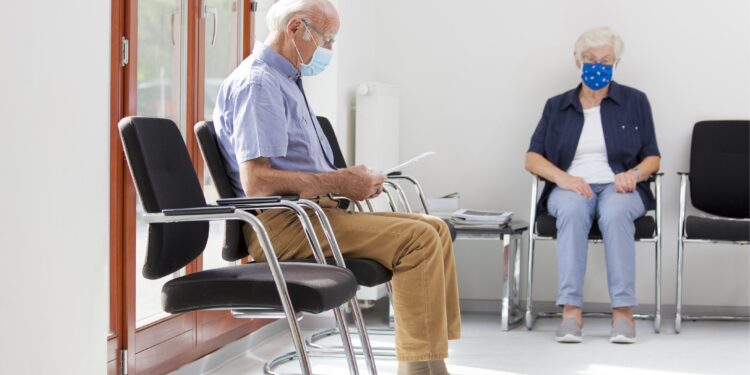 Eine Seniorin und ein Senior mit Mund-Nasen-Bedeckung sitzen in einem Wartezimmer