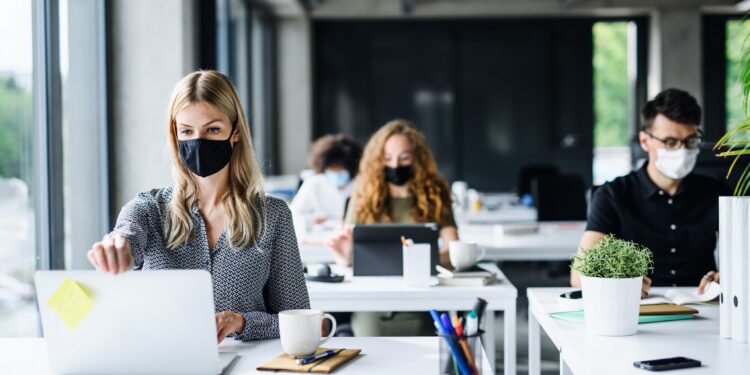 Mehrere Personen sitzen mit Mund-Nasen-Bedeckung vor ihrem Computer in einem Büro