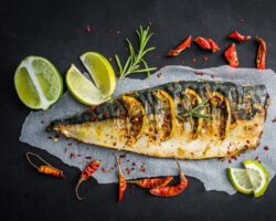 Ein zubereiteter Fisch, umgeben von Limettenscheiben und Gewürzen, liegt auf einer dunklen Oberfläche.