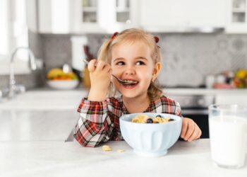 Ein kleines Mädchen, das Frühstück mit Müsli, Milch und Blaubeeren in der Küche isst