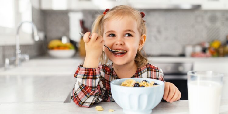 Ein kleines Mädchen, das Frühstück mit Müsli, Milch und Blaubeeren in der Küche isst