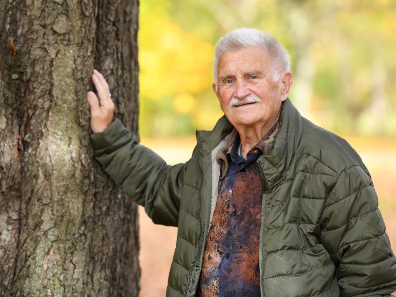 Ein älterer Mann lehnt sich mit der Hand an einen Baum an.