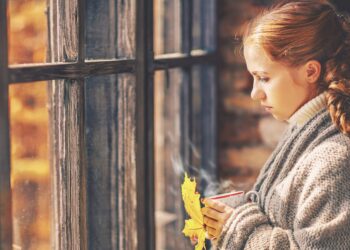 Junge Frau mit einer Tasse in der Hand blickt traurig aus dem Fenster