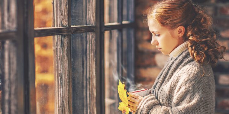 Junge Frau mit einer Tasse in der Hand blickt traurig aus dem Fenster