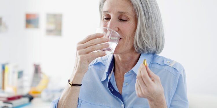 Ältere Frau trinkt aus einem Glas Wasser und hält eine Omega-3-Kapsel